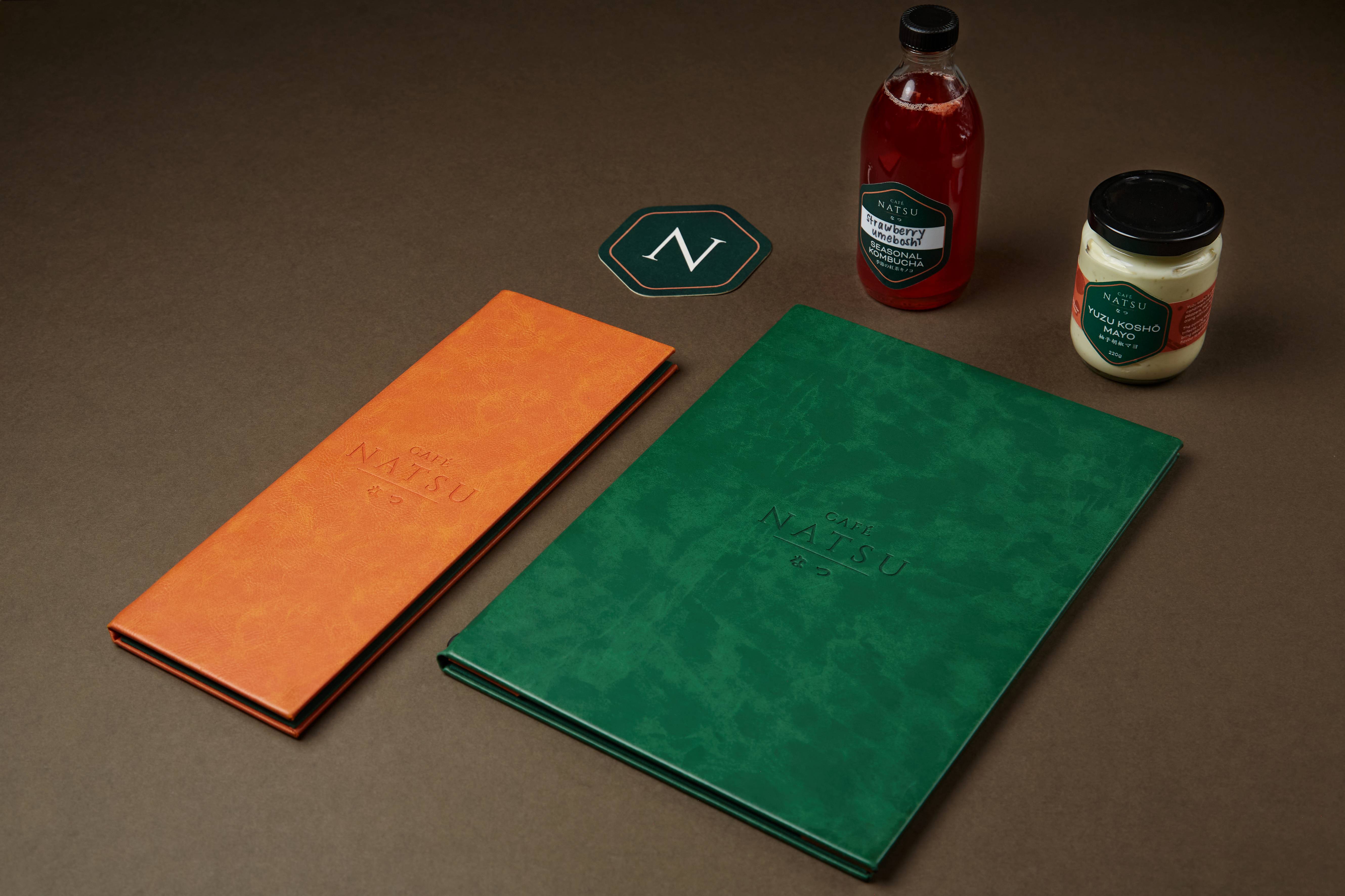 Café Natsu collateral flat-lay on dark brown — coral embossed leather drinks menu, emerald embossed food menu, hexagonal N sticker, strawberry umeboshi kombucha bottle, and yuzu kosho mayo jar