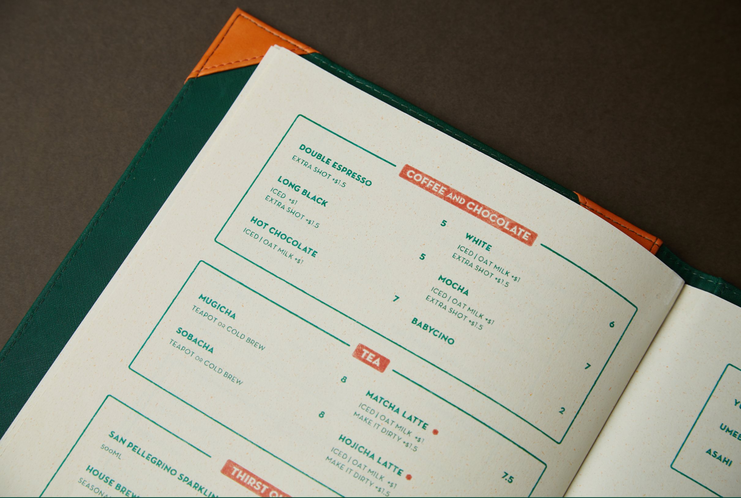 Open Café Natsu menu tilted on a dark surface showing the Coffee and Chocolate and Tea sections in bold green and coral rubber-stamp typography on cream speckled paper
