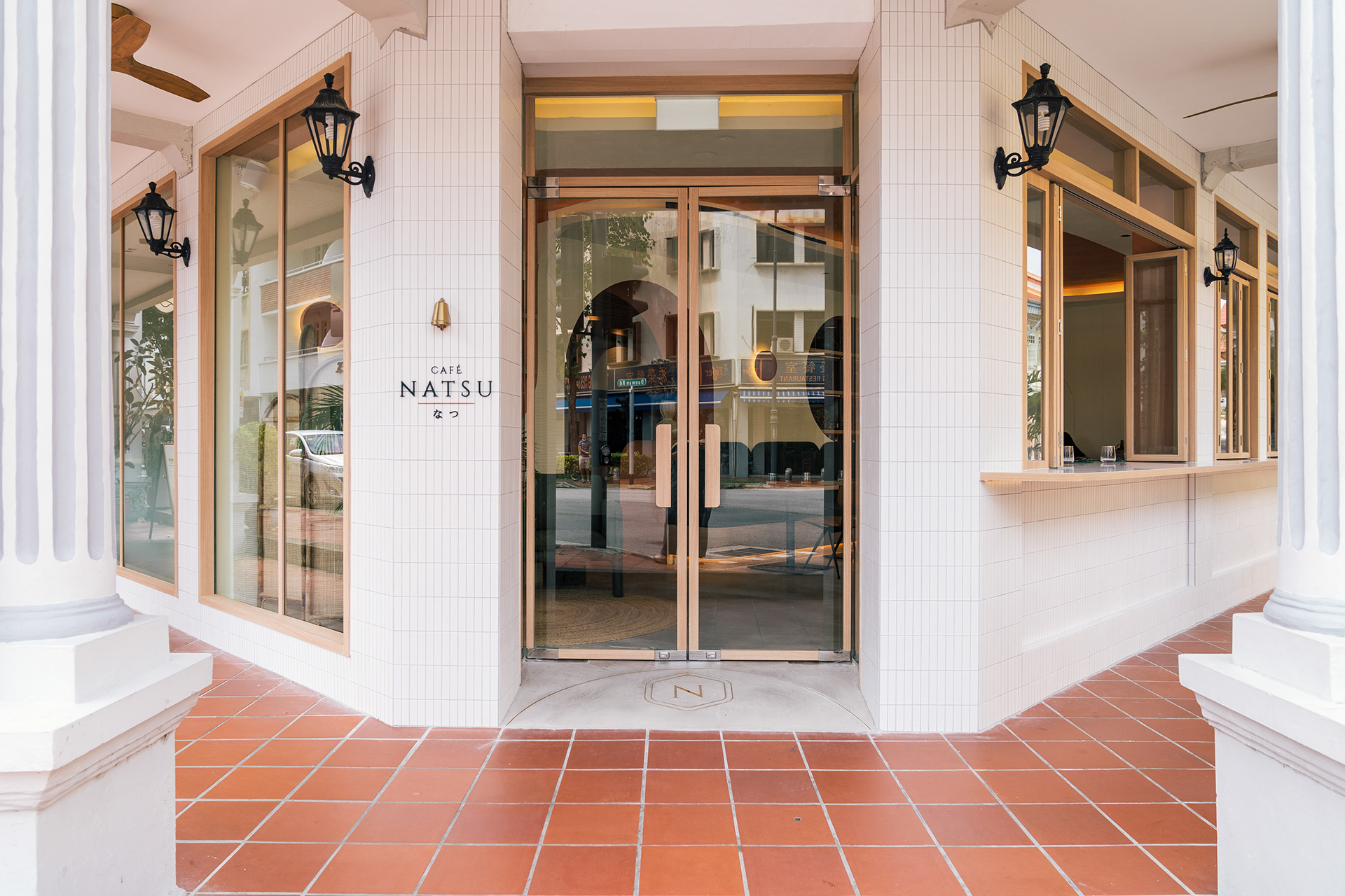 Café Natsu entrance facade — white-tiled shophouse, gold-framed glass doors with CAFÉ NATSU なつ wall signage and terracotta tile forecourt