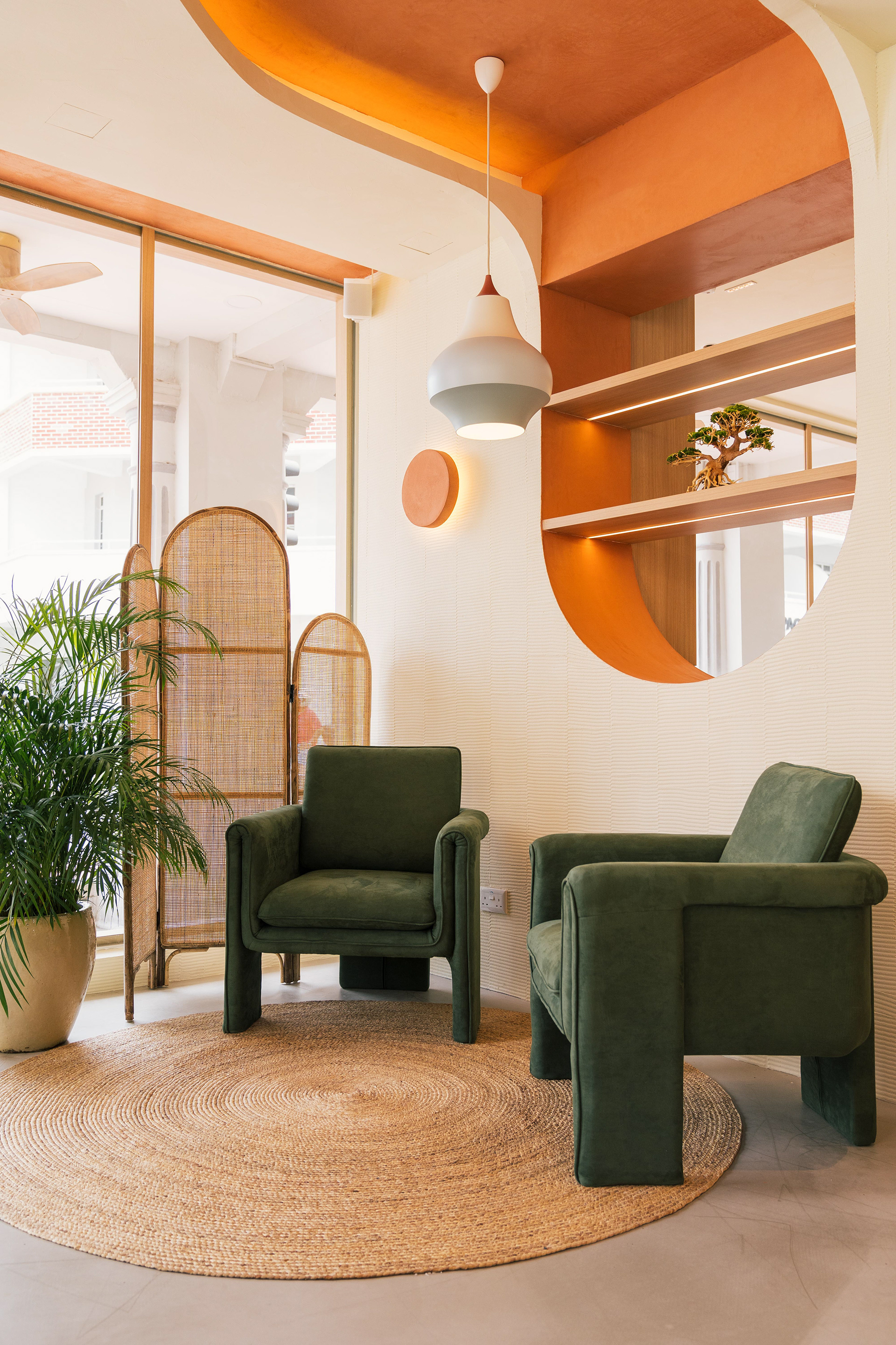 Café Natsu lounge corner — two deep-green armchairs on a round jute rug, rattan room divider, coral orange ceiling cutout, and bonsai on an illuminated niche shelf