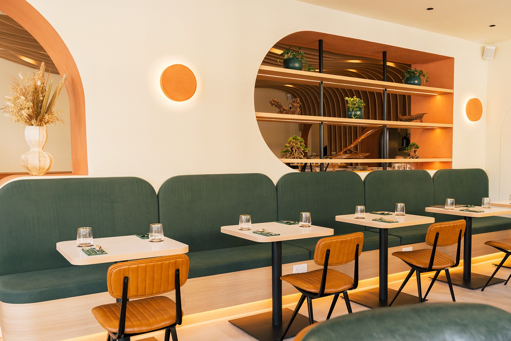 Main dining room — forest-green upholstered booth seating, sand-coloured timber tables, tan leather chairs, large oval orange-lit display shelving, circular disc sconces