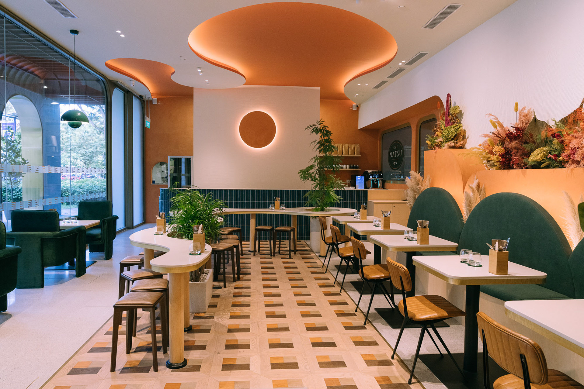 Café Natsu Orchard branch — organic coral ceiling clouds, geometric parquet floor, curved white island counter with green tile, lush plants, and a dried flower installation