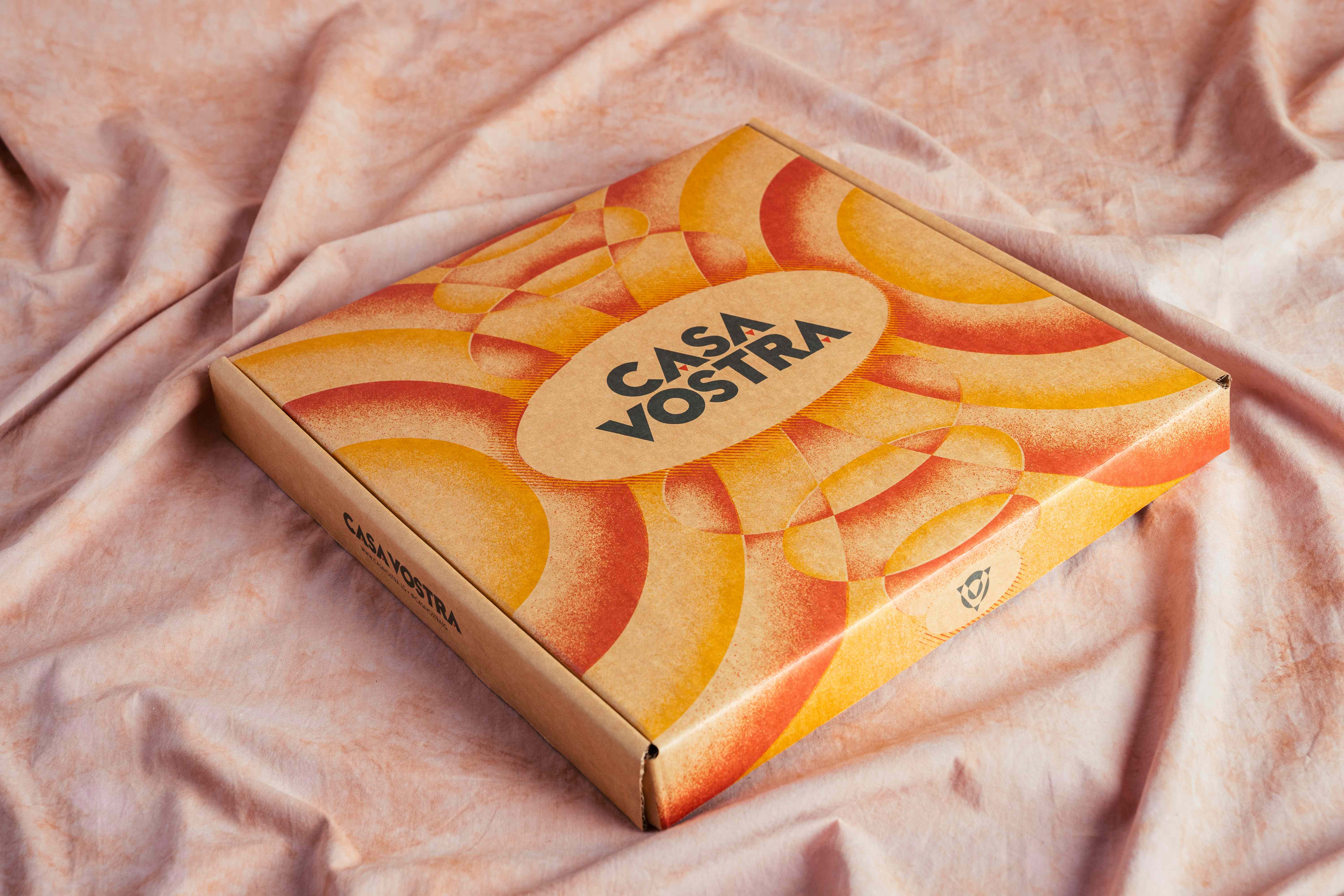 Casa Vostra mailer box with an orange and yellow sunburst pattern and oval logo, resting on pink fabric