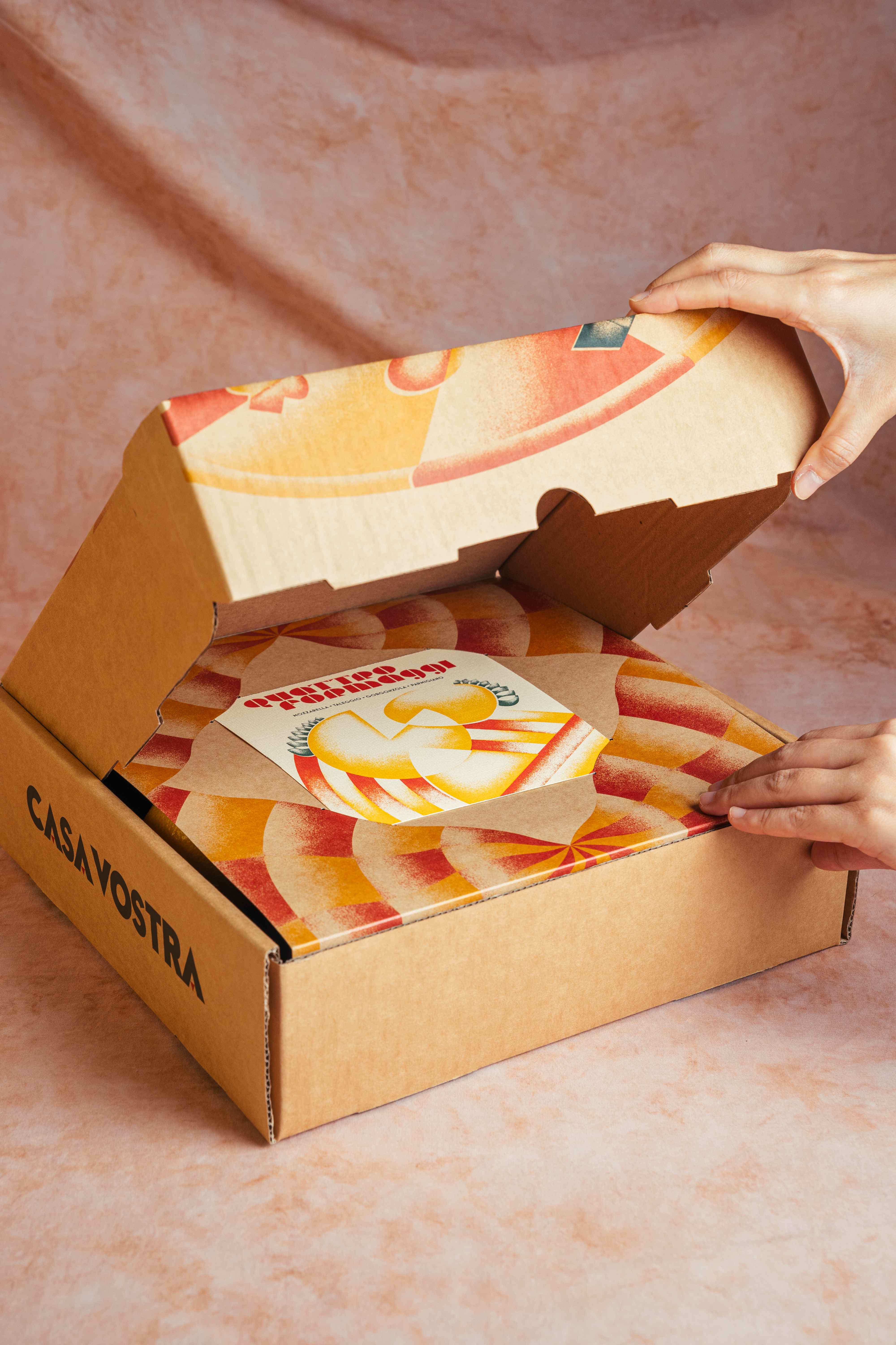 Hands opening a Casa Vostra pizza box, revealing a Quattro Formaggi card and sunburst-printed interior