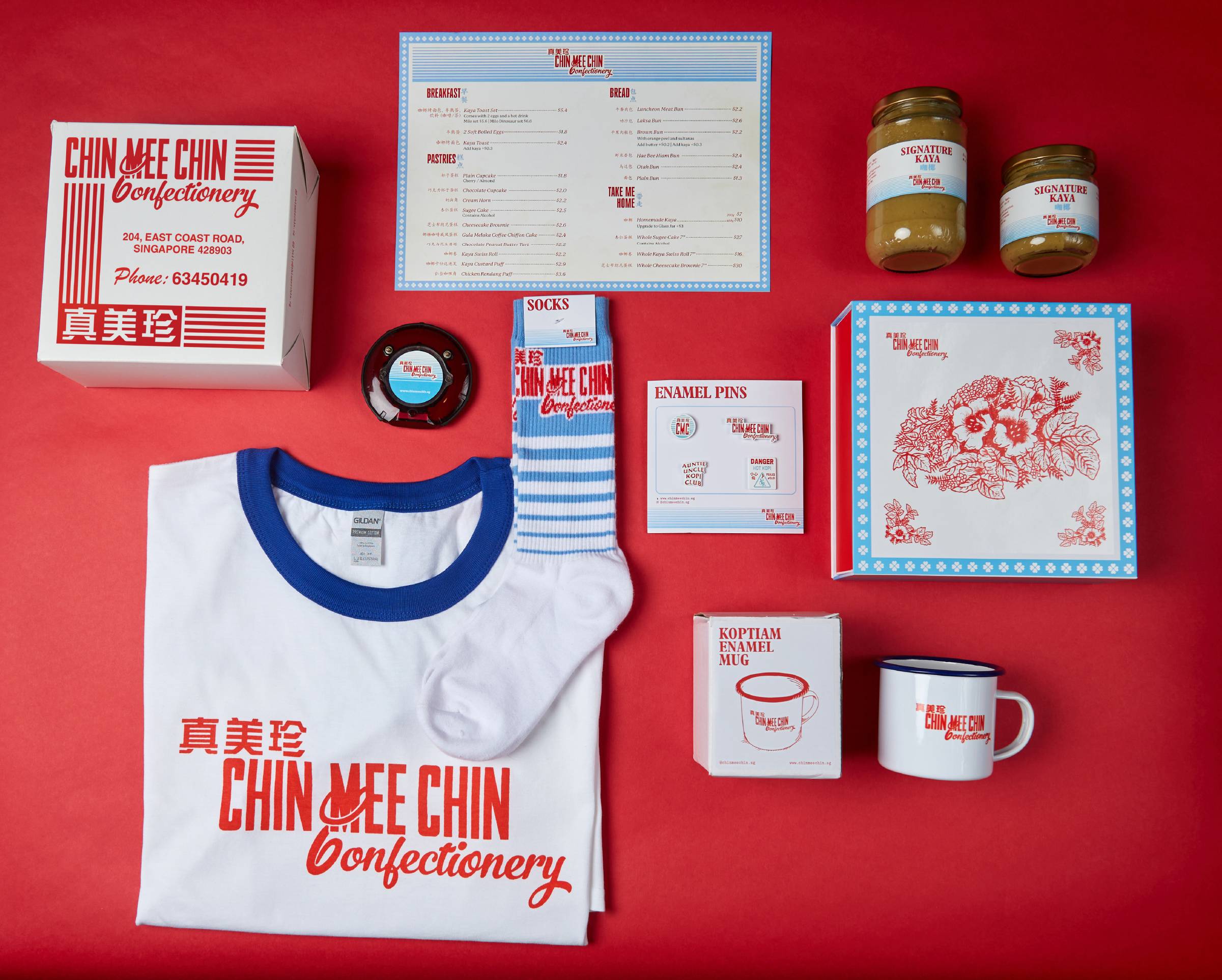 Full CMC merchandise flat-lay on red — t-shirt, socks, enamel pins board, kaya jars, menu, koptiam enamel mug, and floral notepad