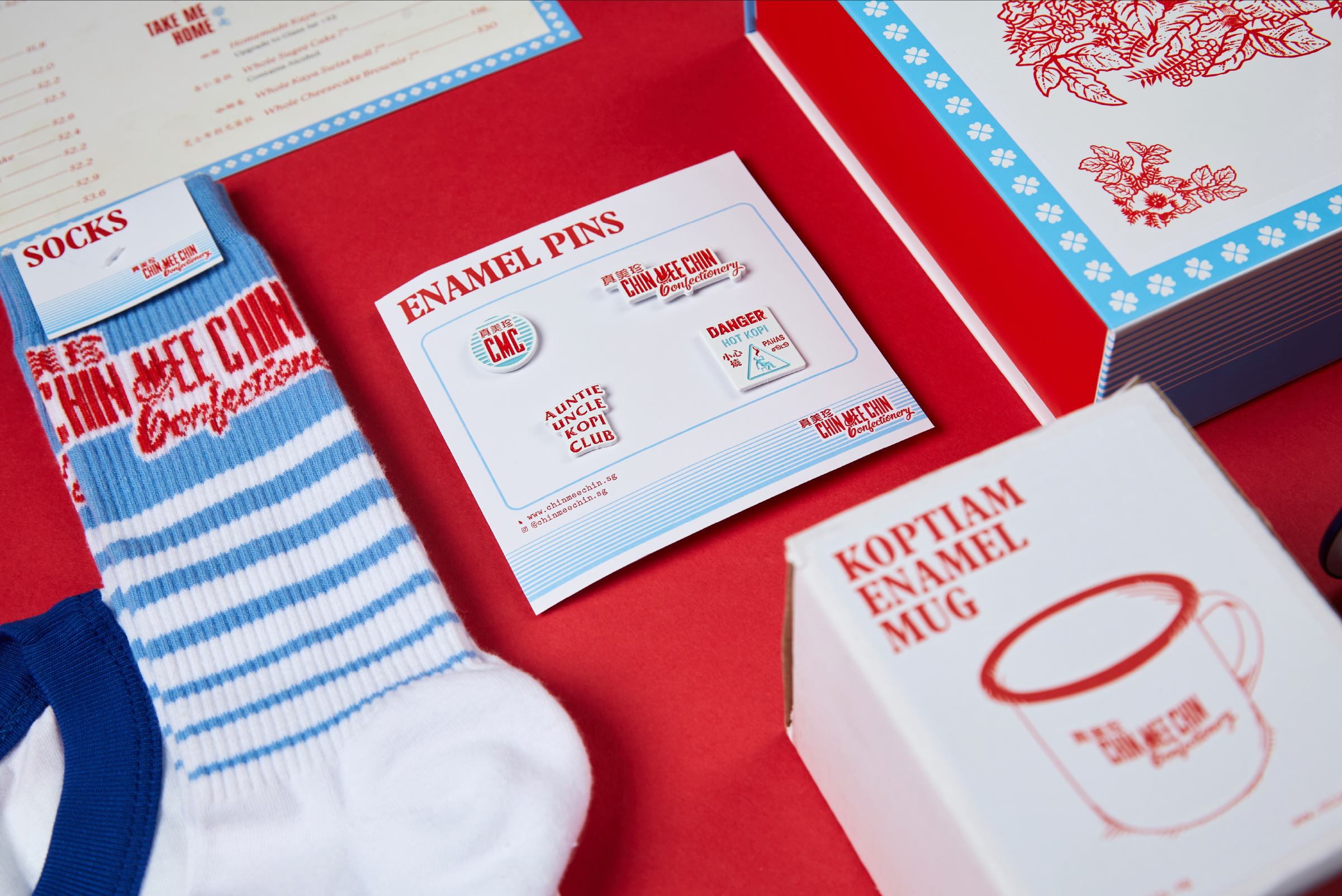 Angled flat-lay on red — enamel pins board, blue-striped socks, koptiam enamel mug box, and floral notepad