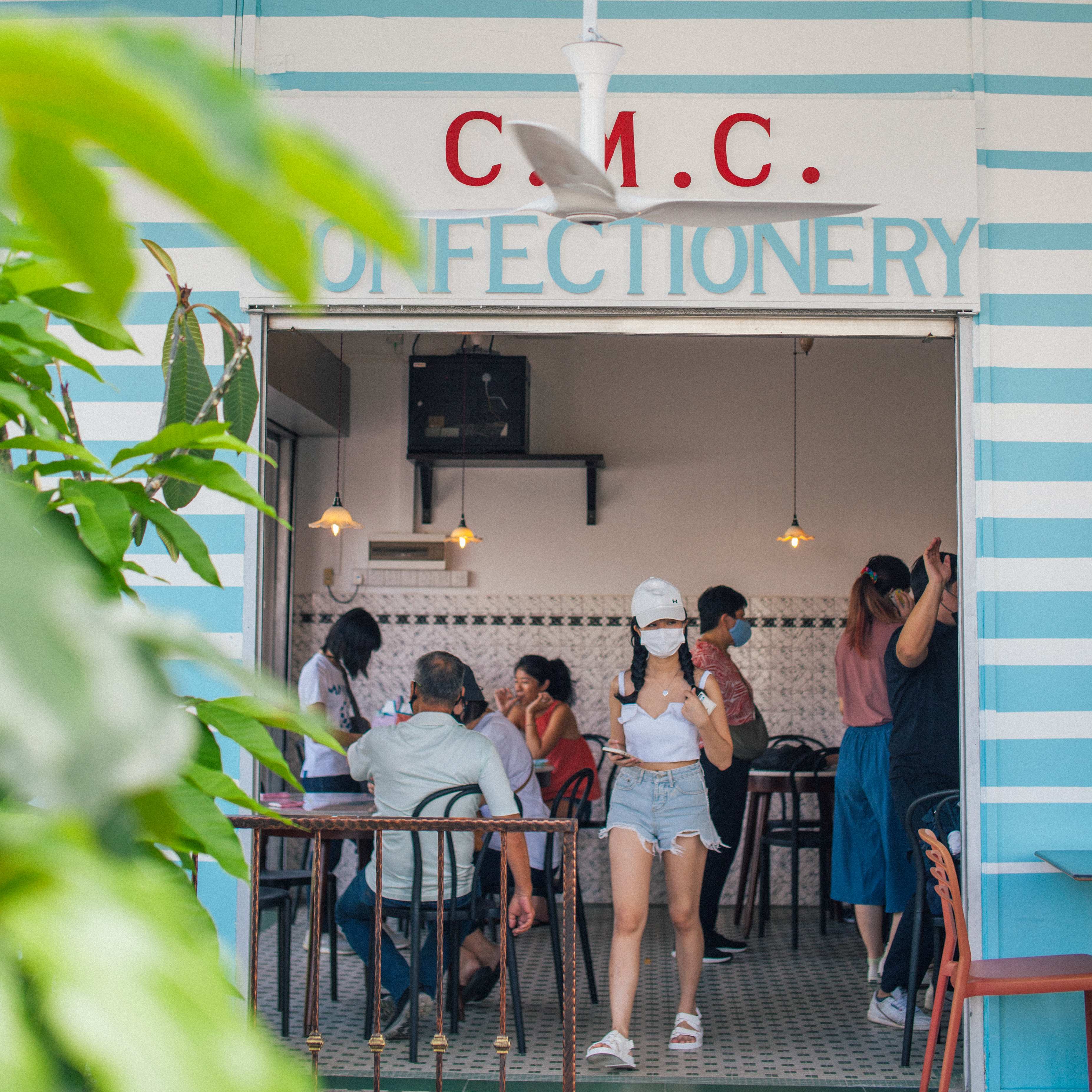 Street-level view framed by tropical foliage, customers visible inside beneath the C.M.C. CONFECTIONERY archway header