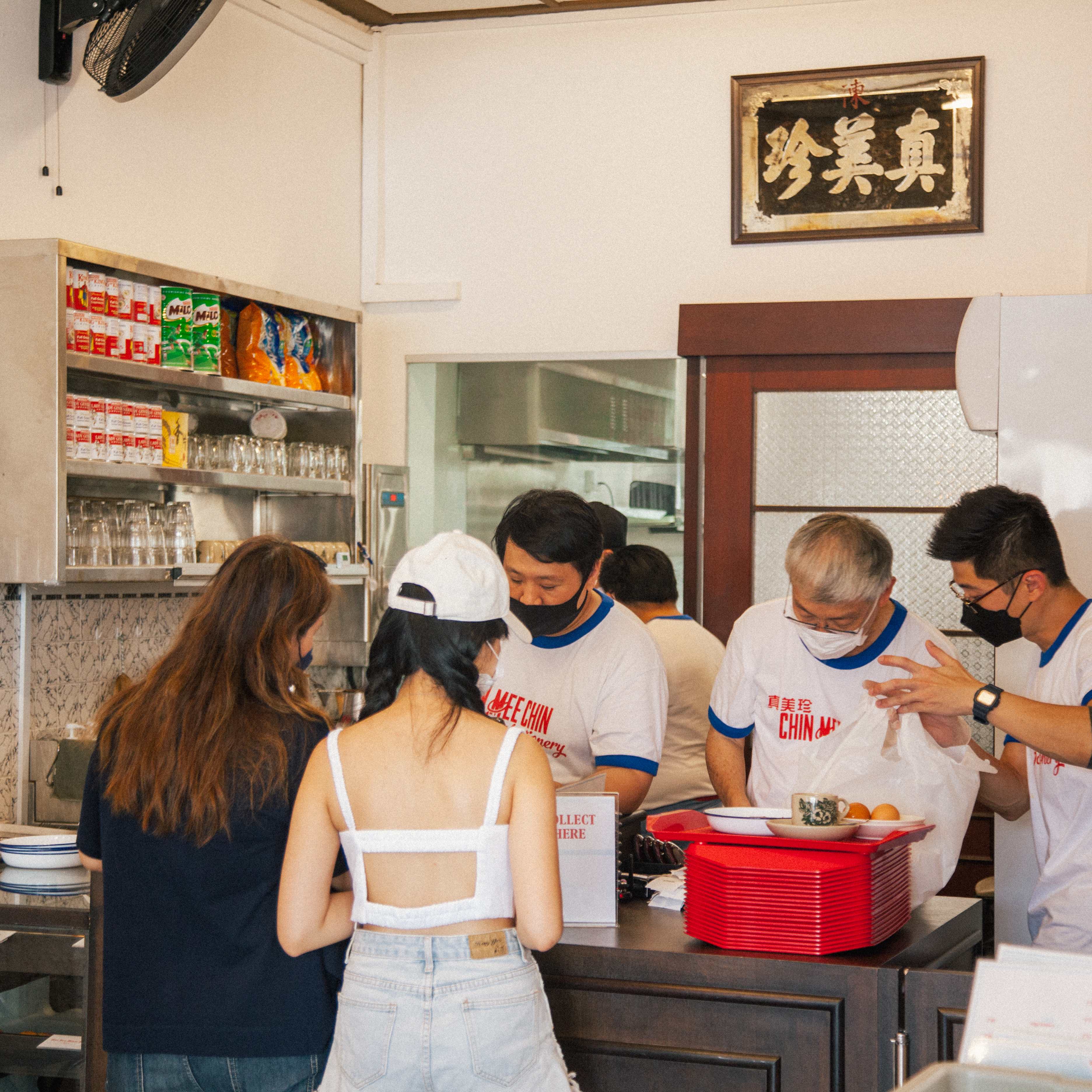 Interior counter scene — staff in branded white-and-blue ringer tees, original Chinese signboard overhead, red trays stacked