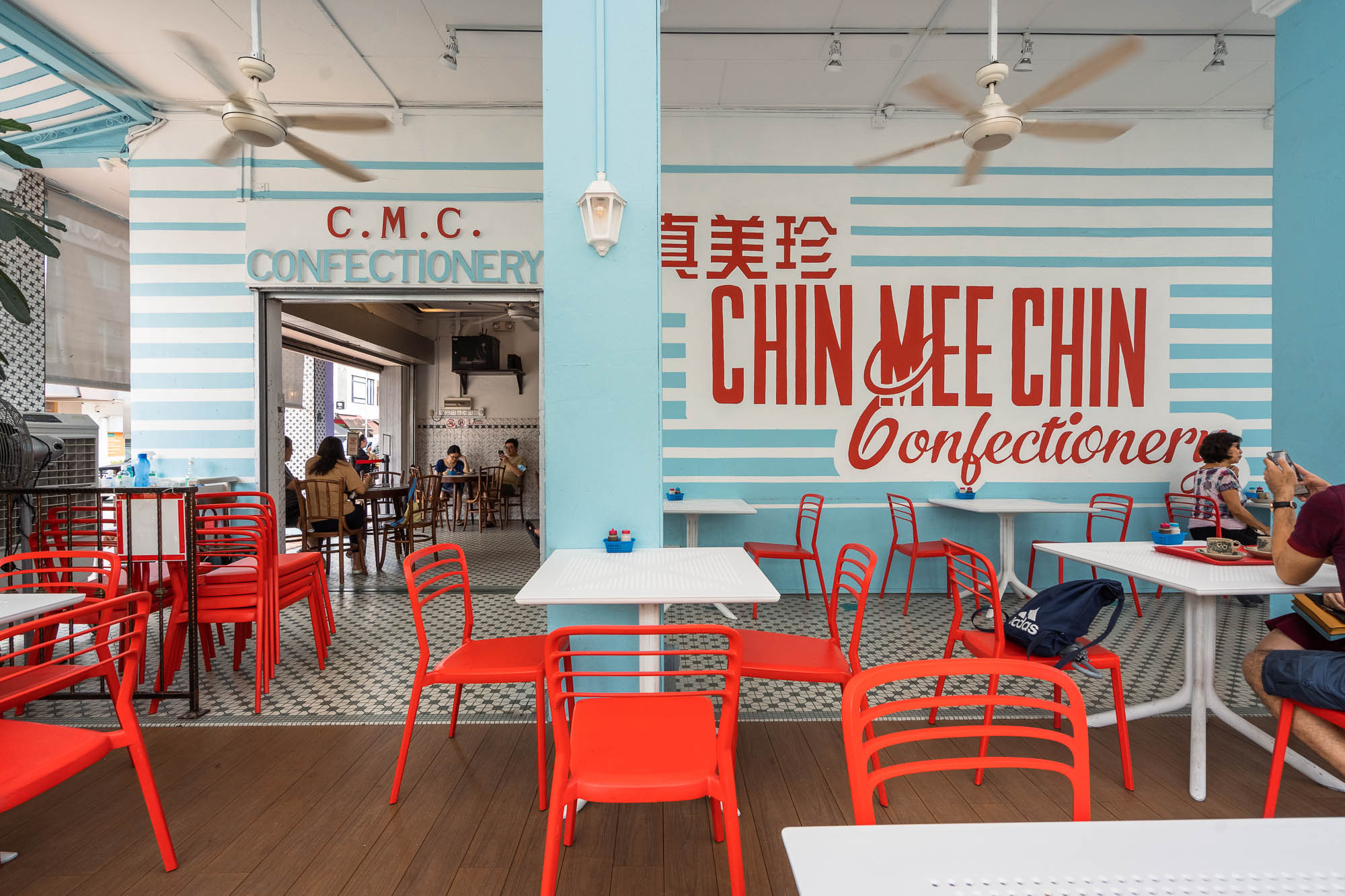 Outdoor terrace with red Tolix chairs, white tables, and the large blue-striped Chin Mee Chin Confectionery wordmark mural