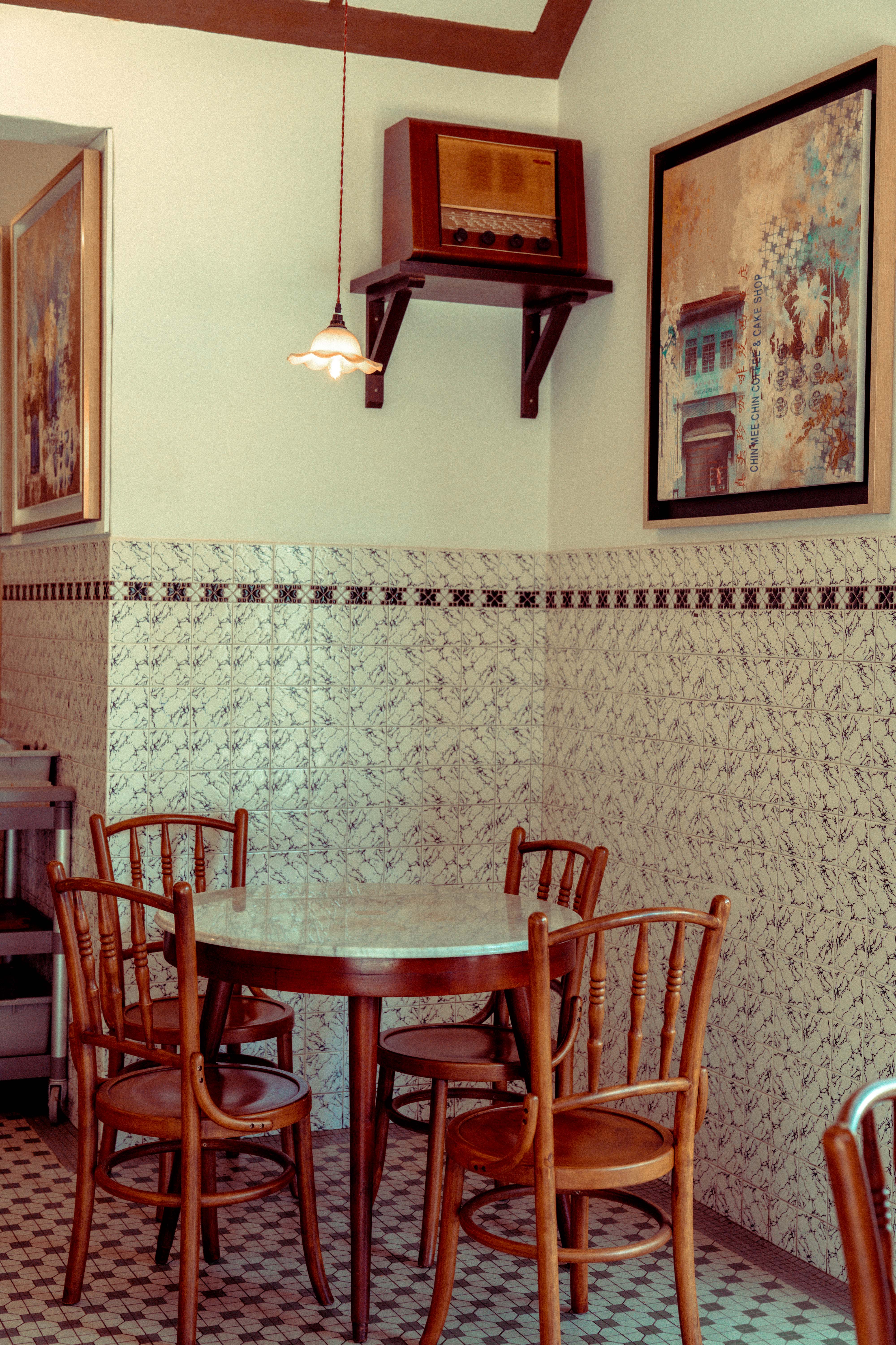 Corner seating with a marble-top round table and wooden bentwood chairs against patterned marble wall tiles, a vintage wooden radio on a shelf, a floral pendant lamp, and a framed heritage shophouse artwork