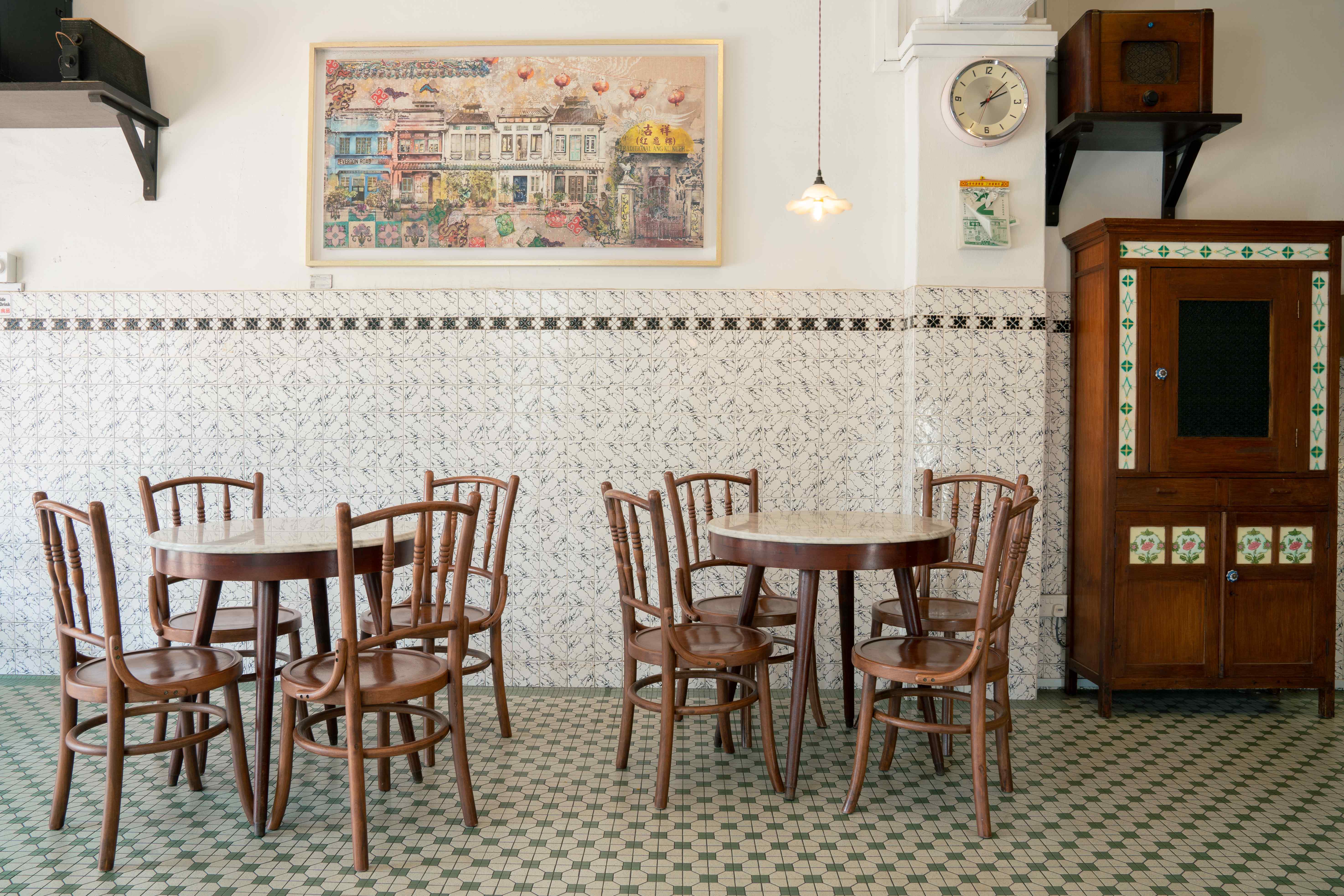 Wide interior view — two marble-top tables with bentwood chairs beneath a large framed Everton Road shophouse painting, a wall clock, a vintage radio on a shelf, and a wooden cabinet with green-diamond tiled panels