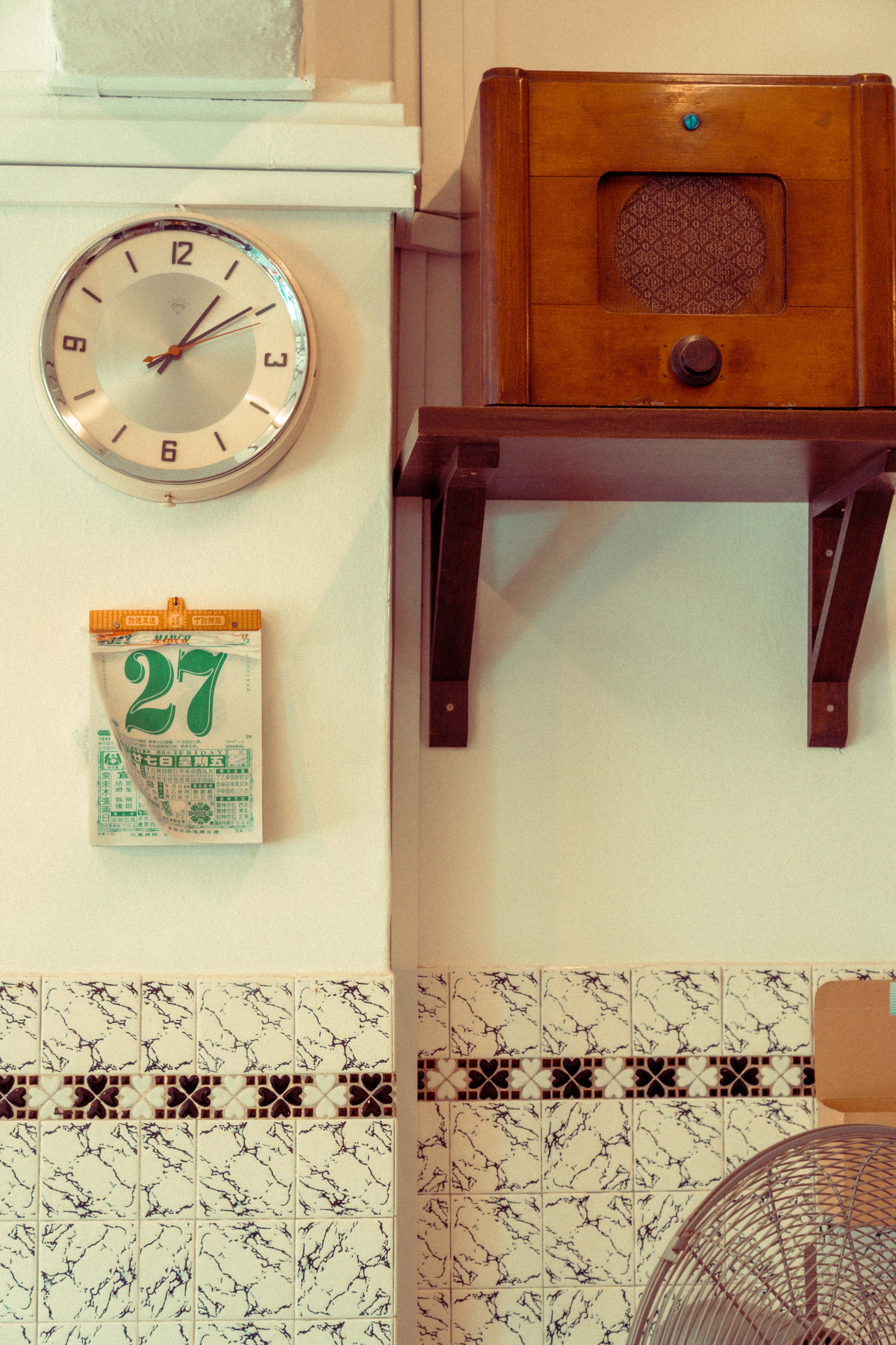 Wall detail — a chrome-rimmed clock, a green '27' tear-off calendar, and a wooden vintage radio on a bracket shelf above marbled tiles with a heart-motif border