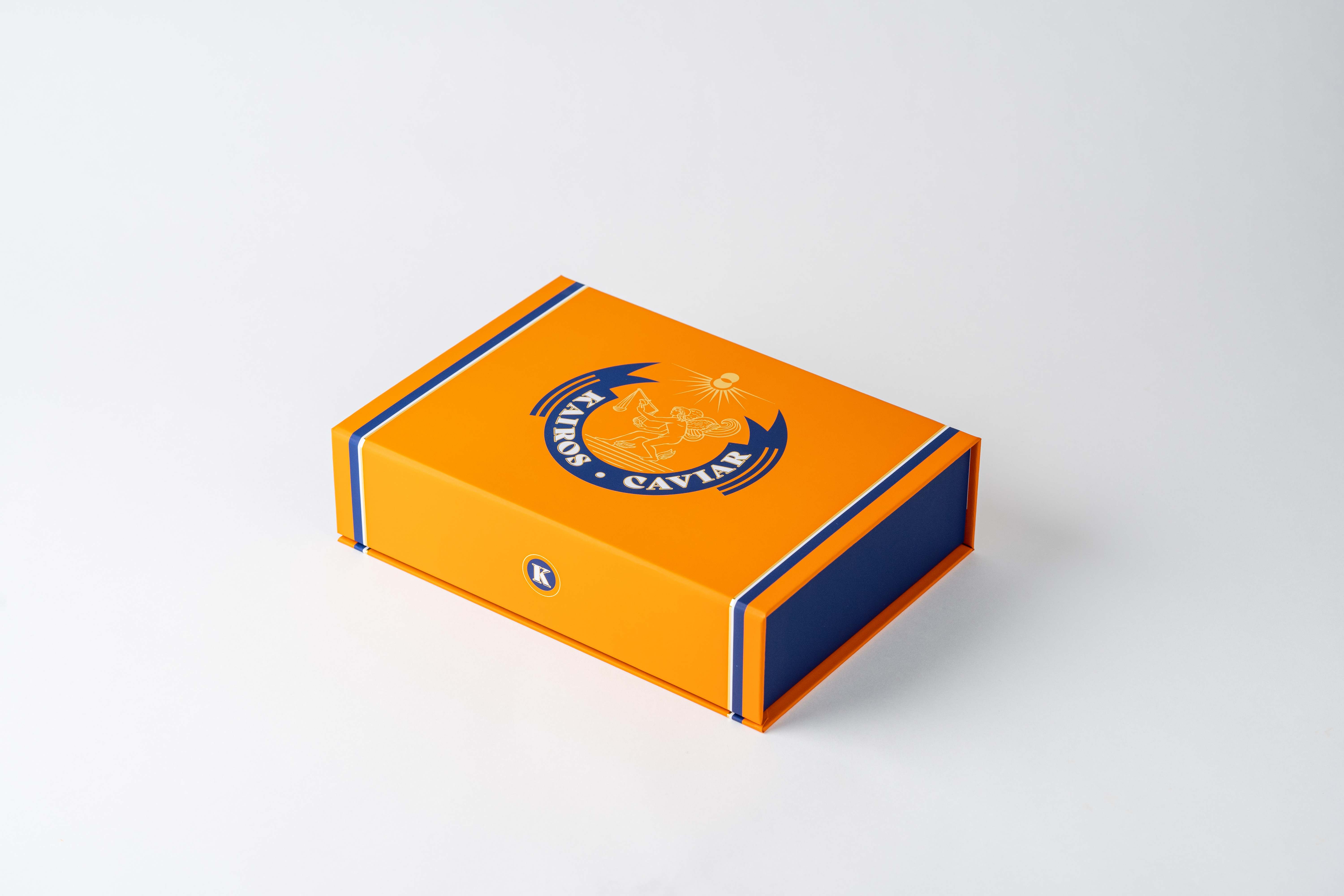 Kairos Caviar magnetic gift box in bright orange with a navy stripe and the winged Kairos crest, closed on a white backdrop