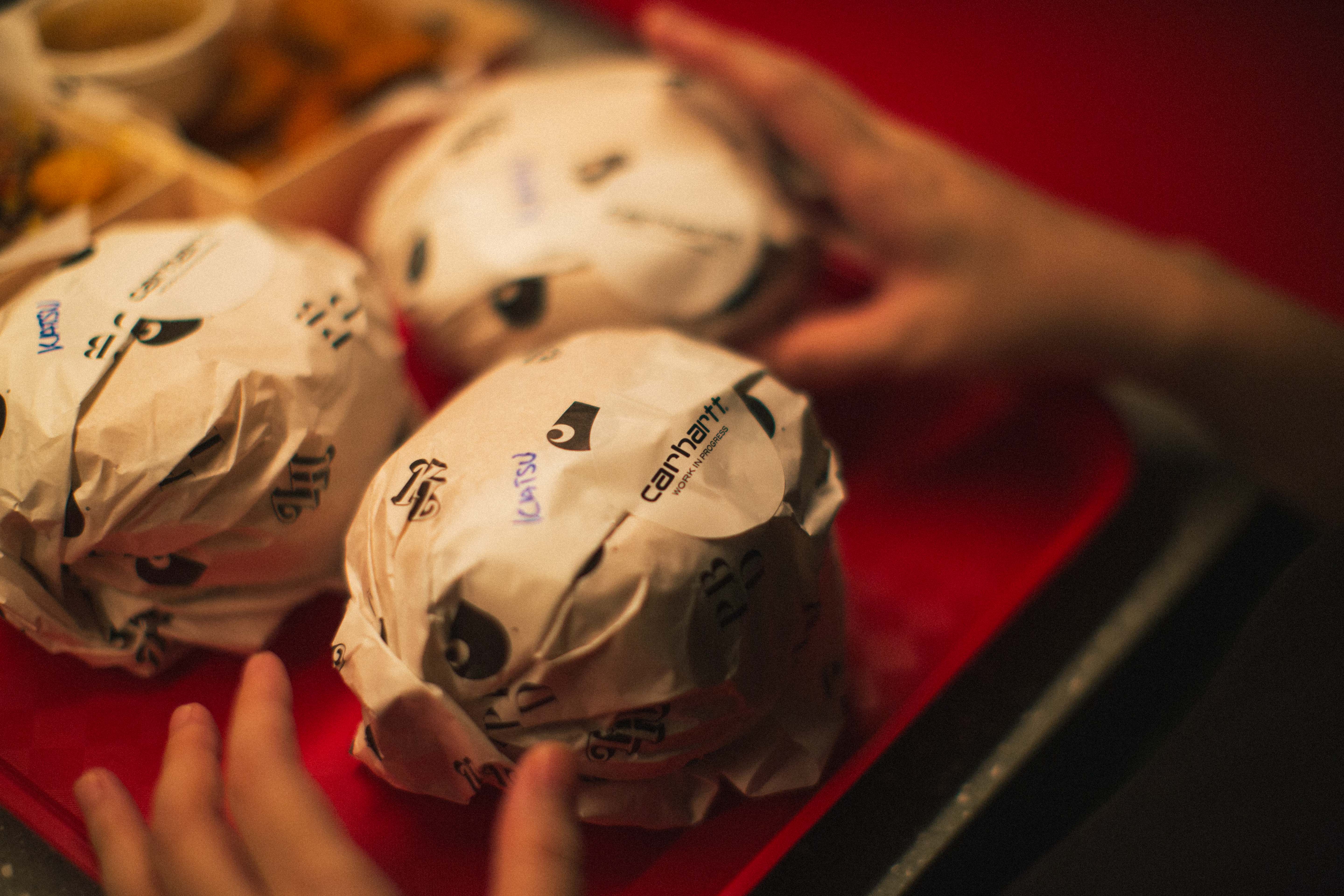 Close-up of burgers wrapped in Love Handle branded paper with Carhartt WIP Katsu stickers on a red tray, hands reaching in