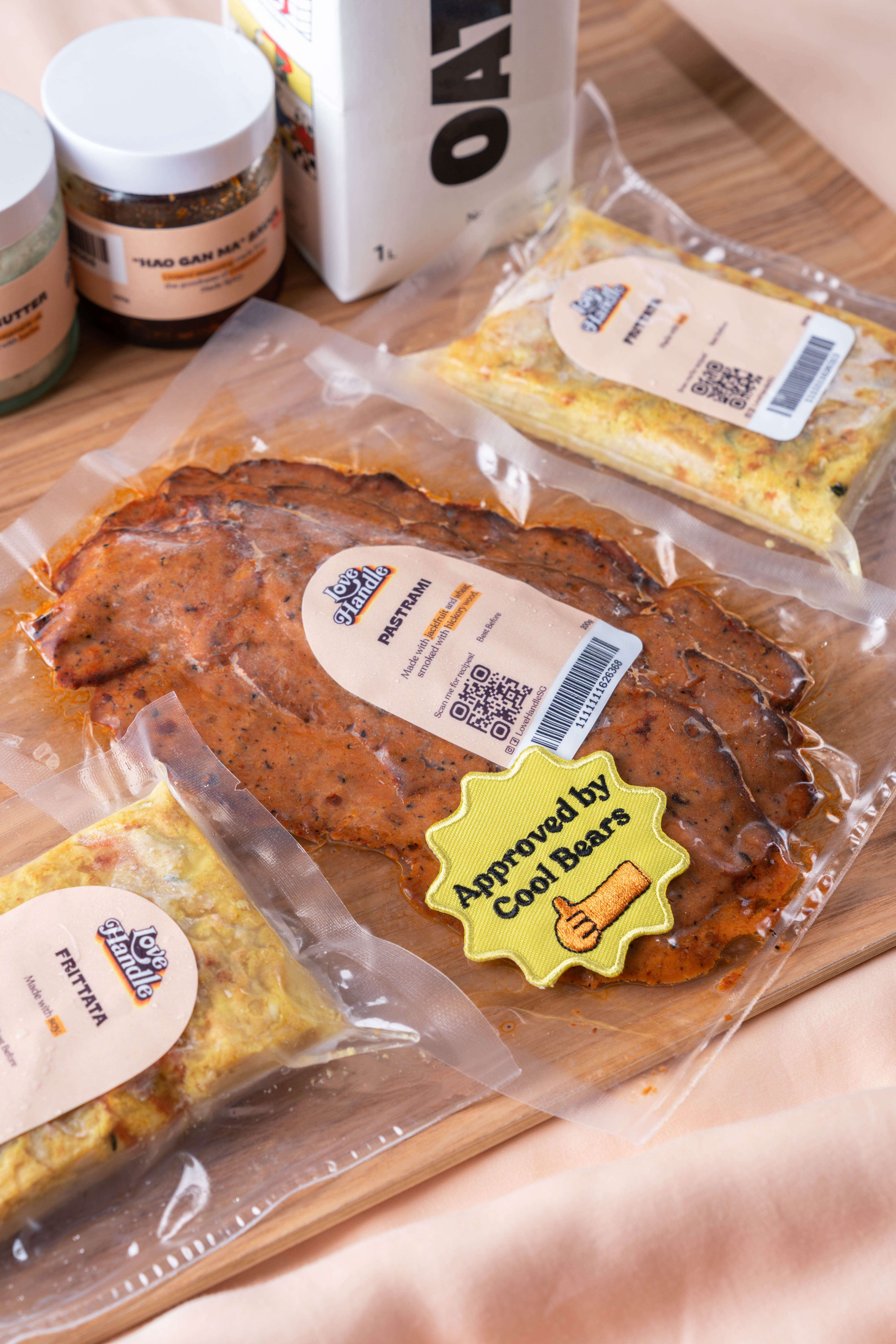 Love Handle products on a wooden board — Pastrami and Frittata vacuum packs, sauce jars and an Approved by Cool Bears embroidered patch