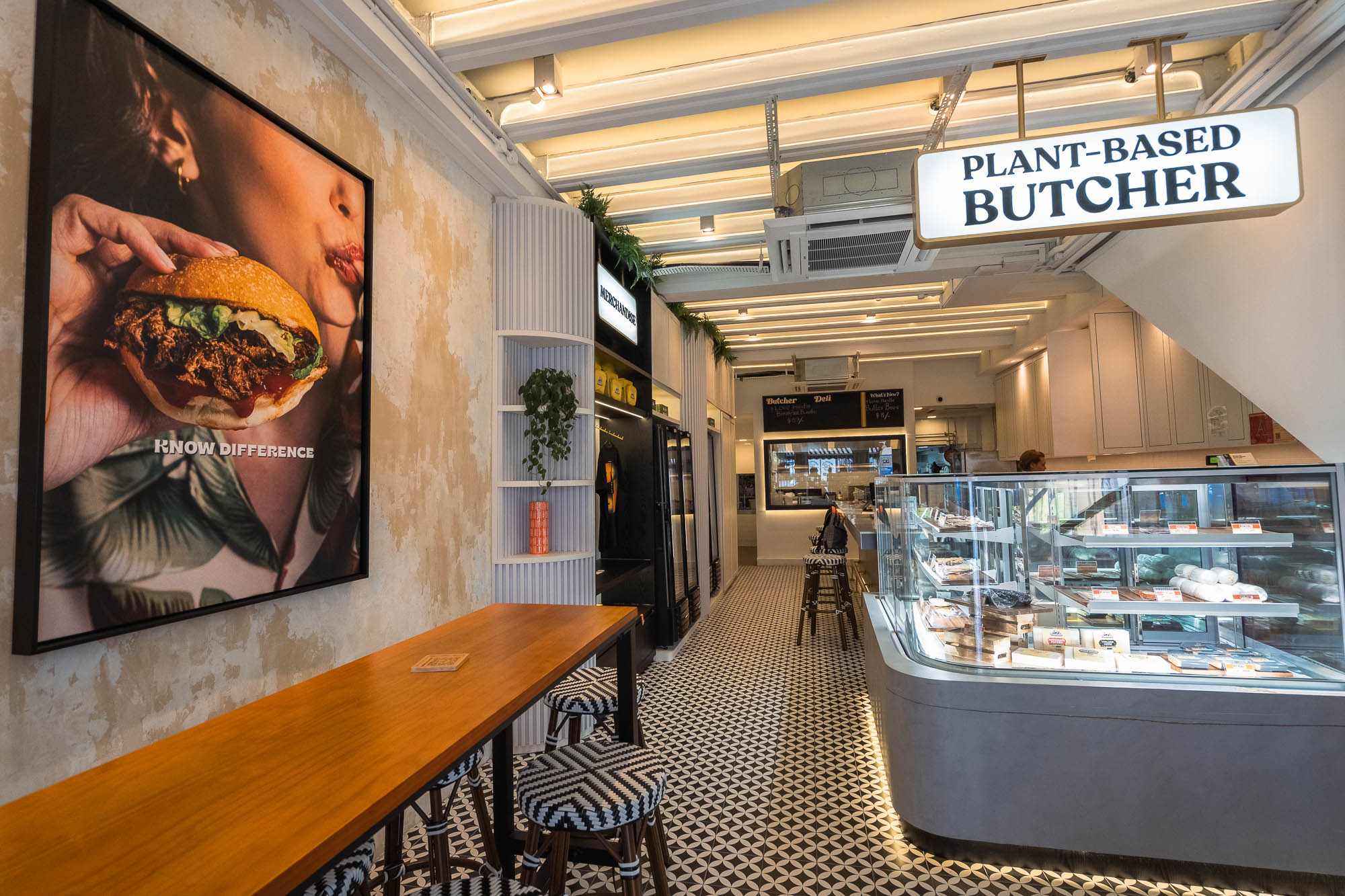 Love Handle Plant-Based Butcher corridor — chilled deli display case, a KNOW DIFFERENCE burger poster on textured plaster, black-and-white patterned floor tiles and bar seating