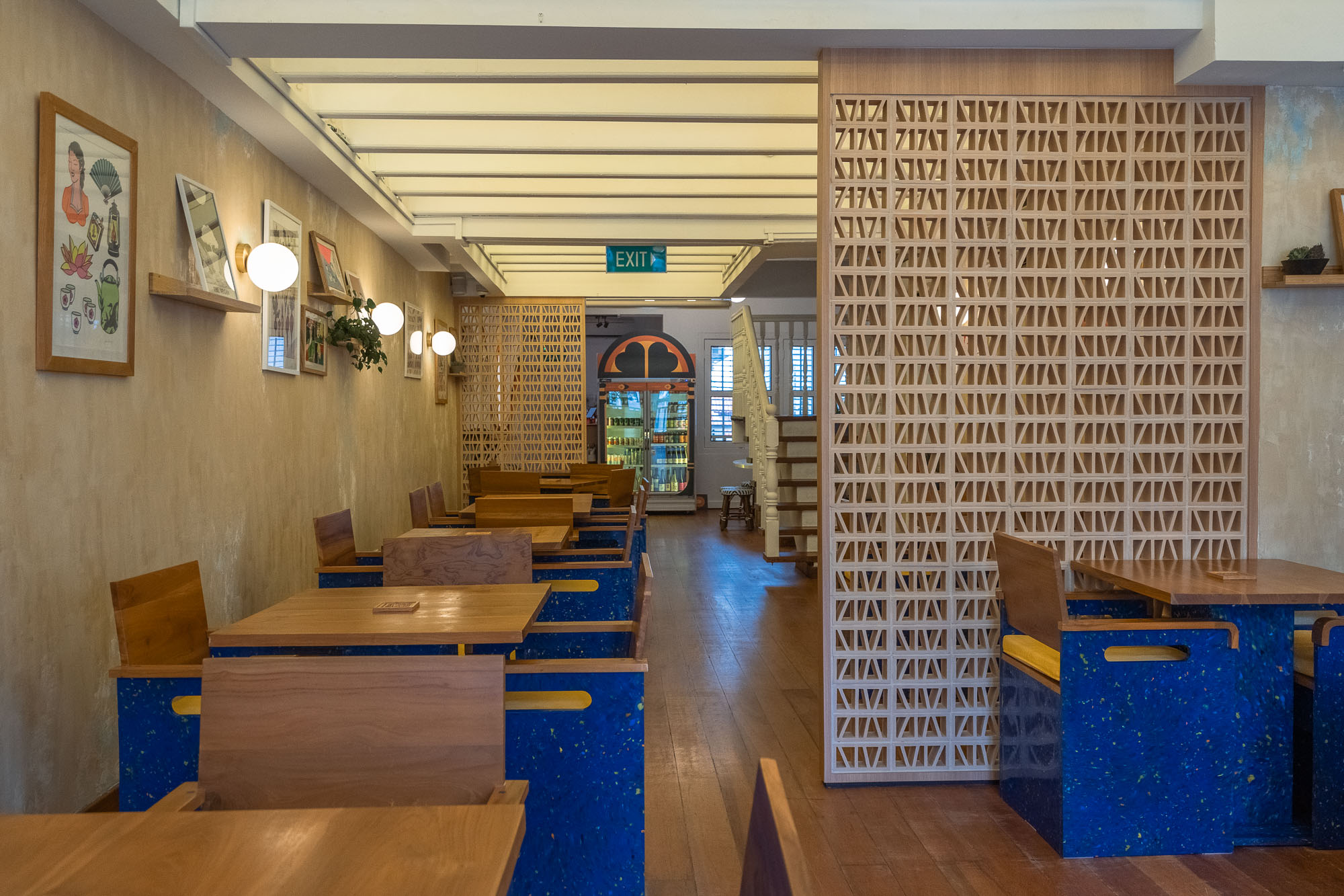 Ground-floor dining corridor — blue recycled-plastic benches, timber tables, a breeze-block screen divider, gallery wall of framed prints and a drinks fridge at the rear