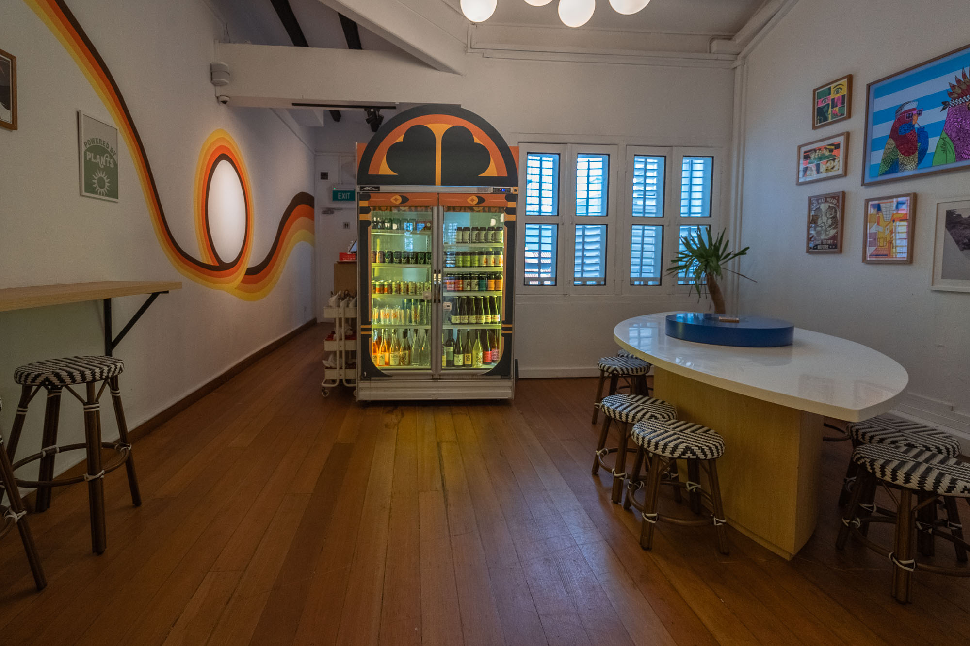 Retro lounge area — rainbow swoosh wall mural, arched graphic drinks fridge stocked with craft brews, an oval bar with patterned rattan stools and a gallery wall