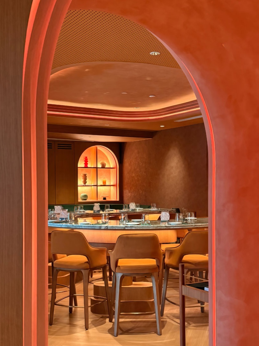 View through a terracotta arch toward the curved marble bar, mustard stools, and a glowing arched display niche