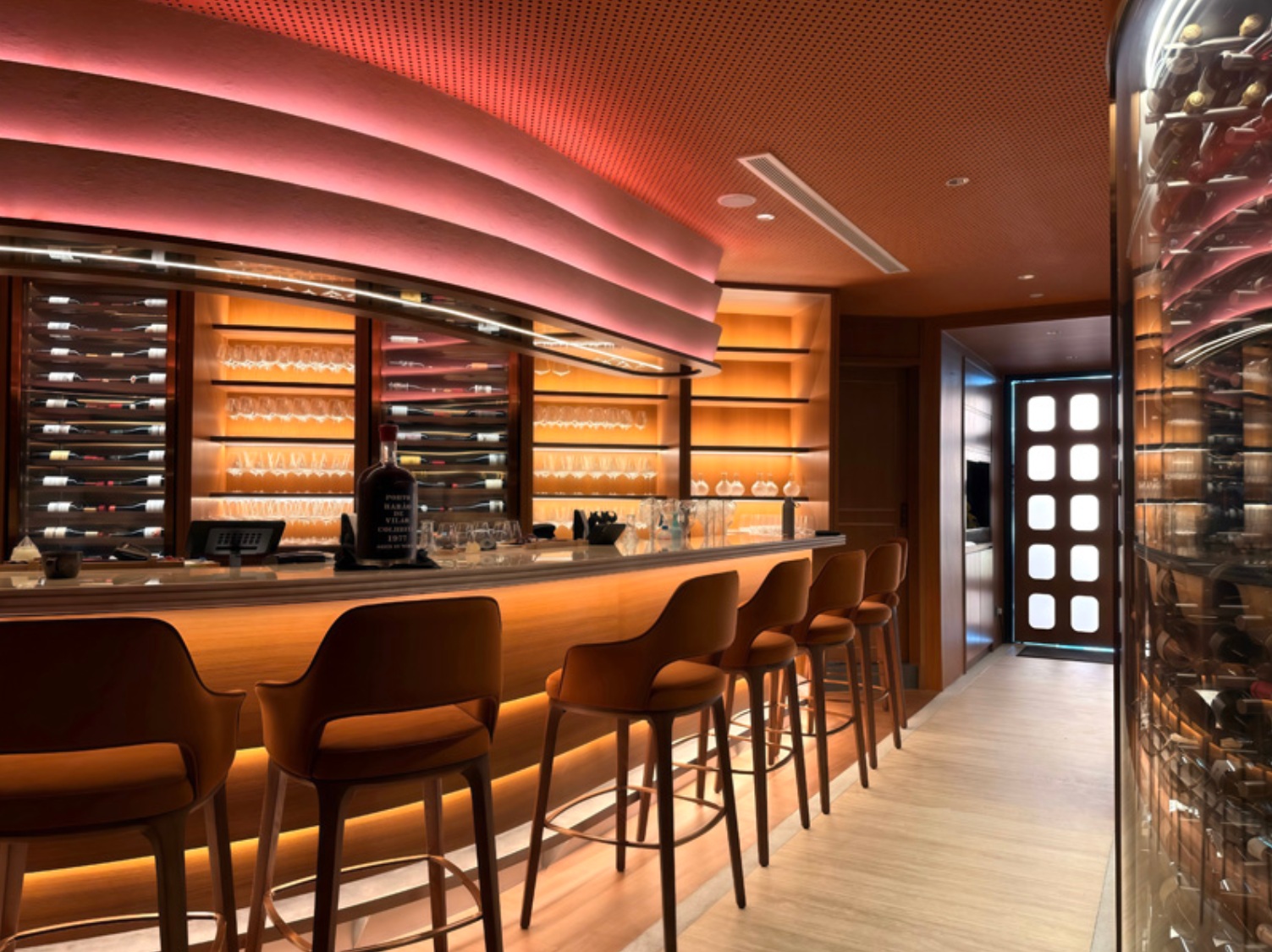 Curved bar with backlit wine display, mustard leather stools, a pink-lit undulating ceiling, and a cylindrical wine fridge
