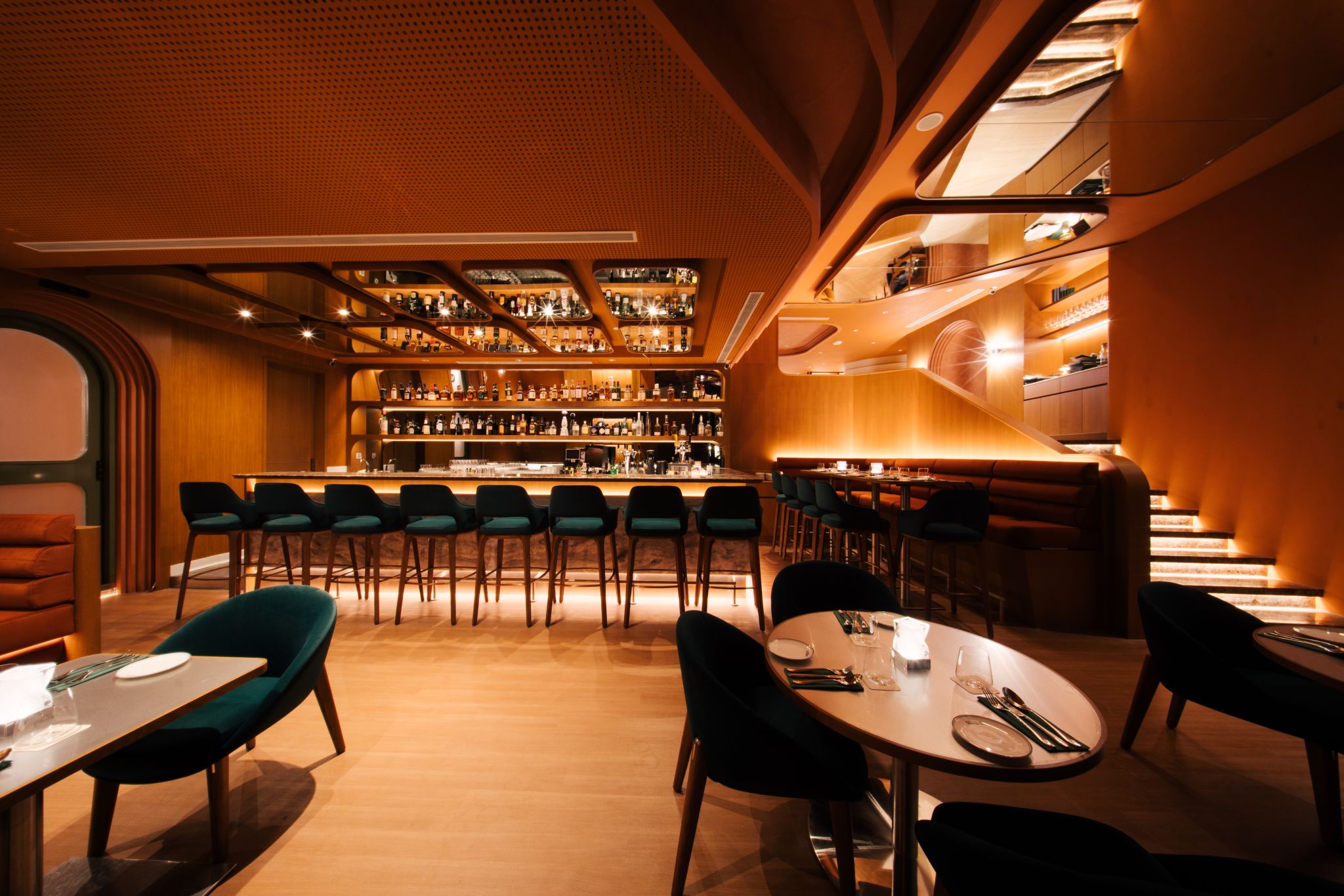 Wide bar room with backlit bottle shelving, green velvet stools, perforated ceiling, and stepped seating