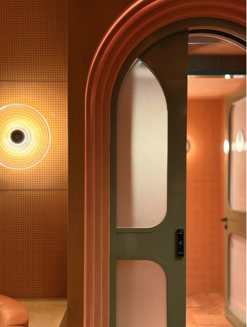Detail of an arched green reeded-glass door framed by a terracotta arch with a circular sconce on a perforated wood wall