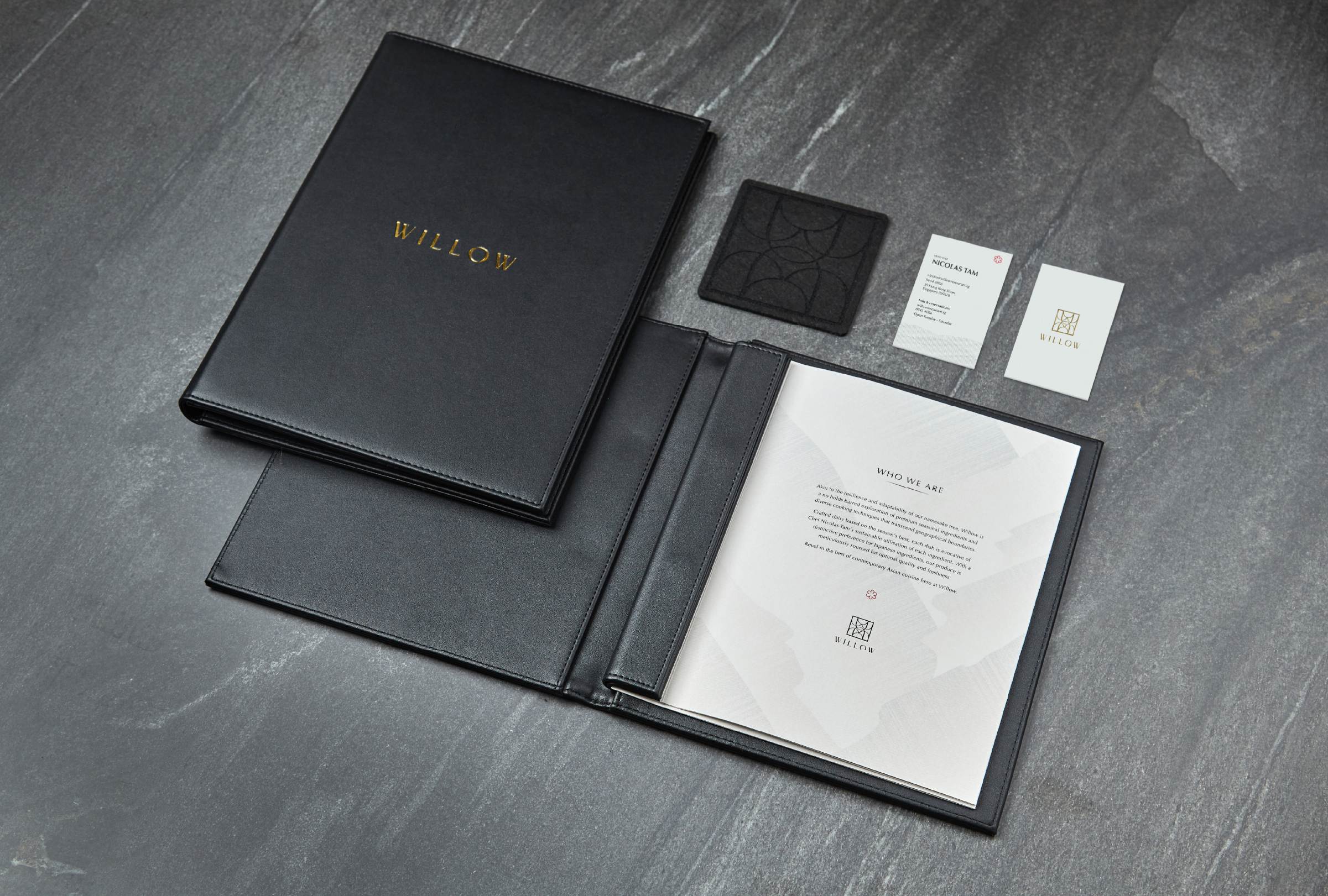 Willow collateral on dark slate — black padded leather menu portfolio with gold foil WILLOW stamp, open to a Who We Are inside spread; business cards and embossed geometric coaster alongside
