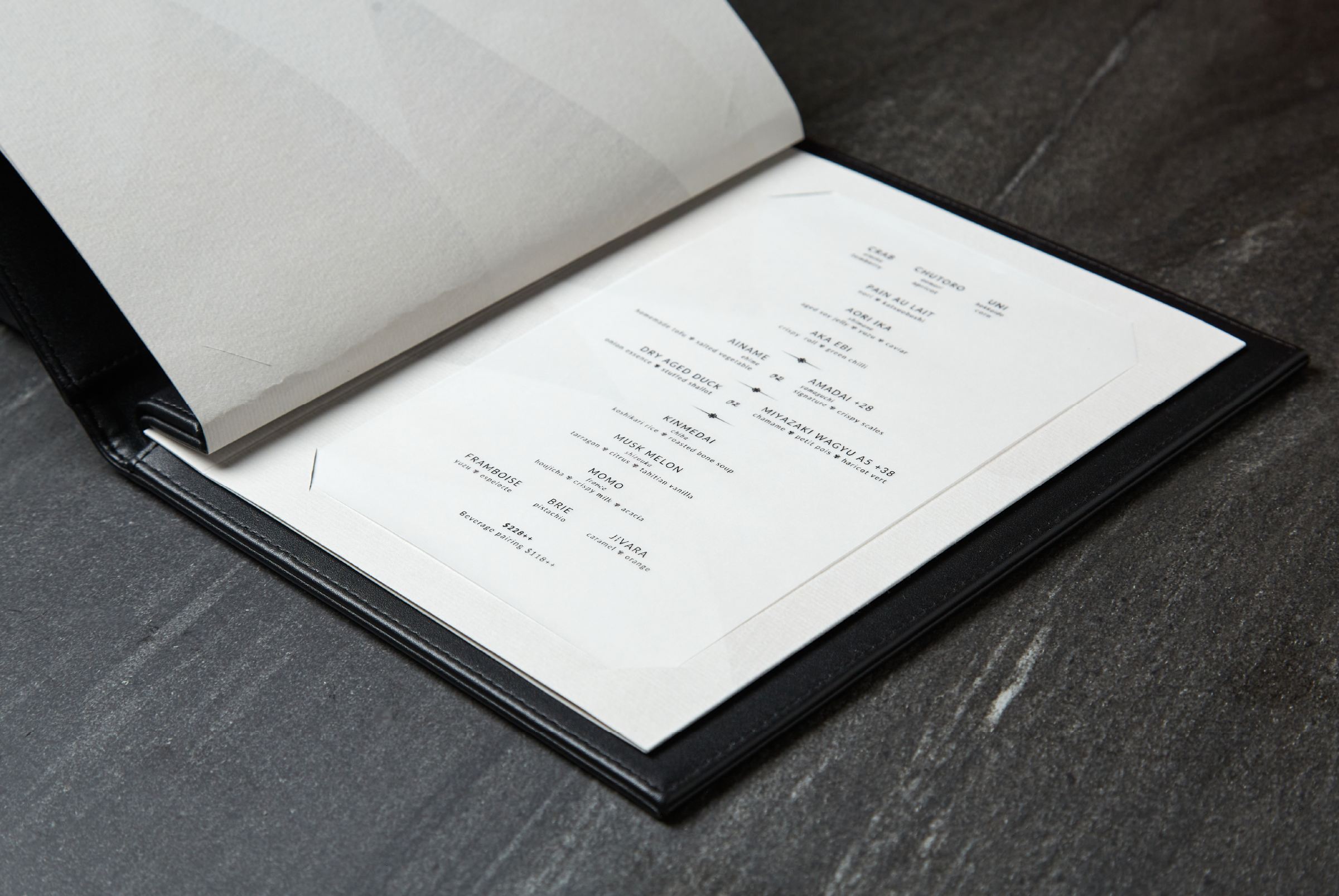 Open Willow tasting menu tilted on dark slate — interior page with delicate scattered course listing in fine black Optima on cream paper with ghost watermark logo pattern