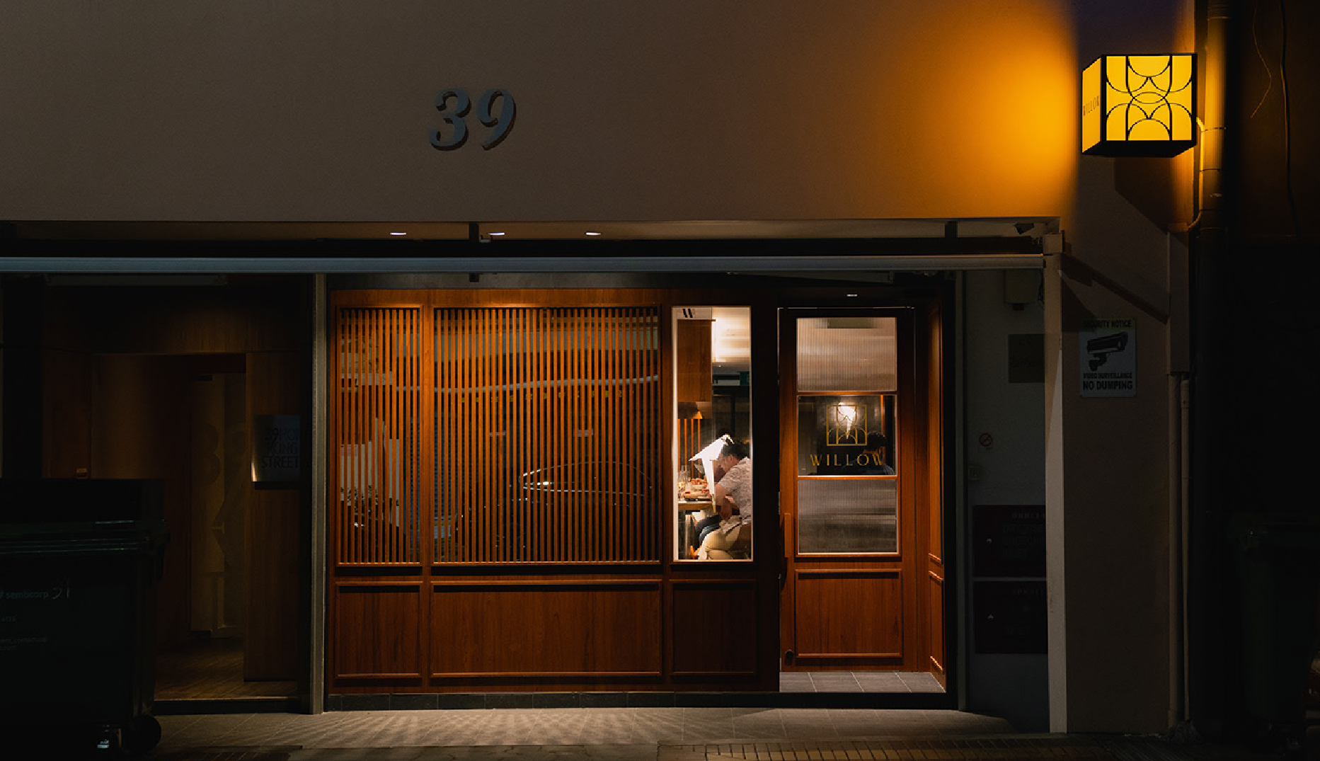Willow restaurant exterior at night — warm amber timber-slat facade at 39 Hong Kong Street, glass entry door with gold WILLOW badge mark, custom lantern sconce casting a halo glow