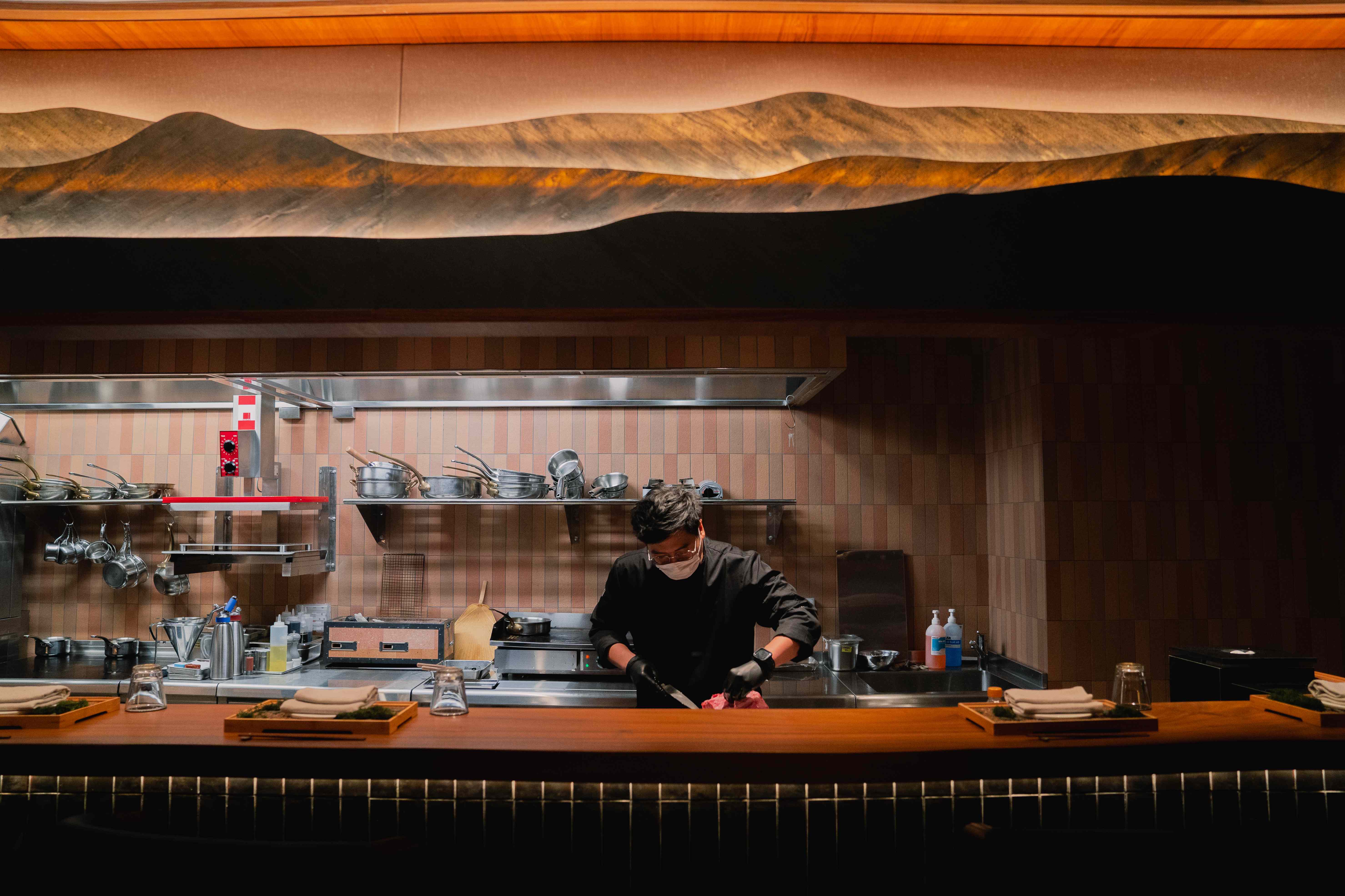 Willow open kitchen — chef in black preparing at the pass, pink-terracotta mosaic tile splashback, undulating hand-sculpted mountain-form sculptural canopy in warm amber above
