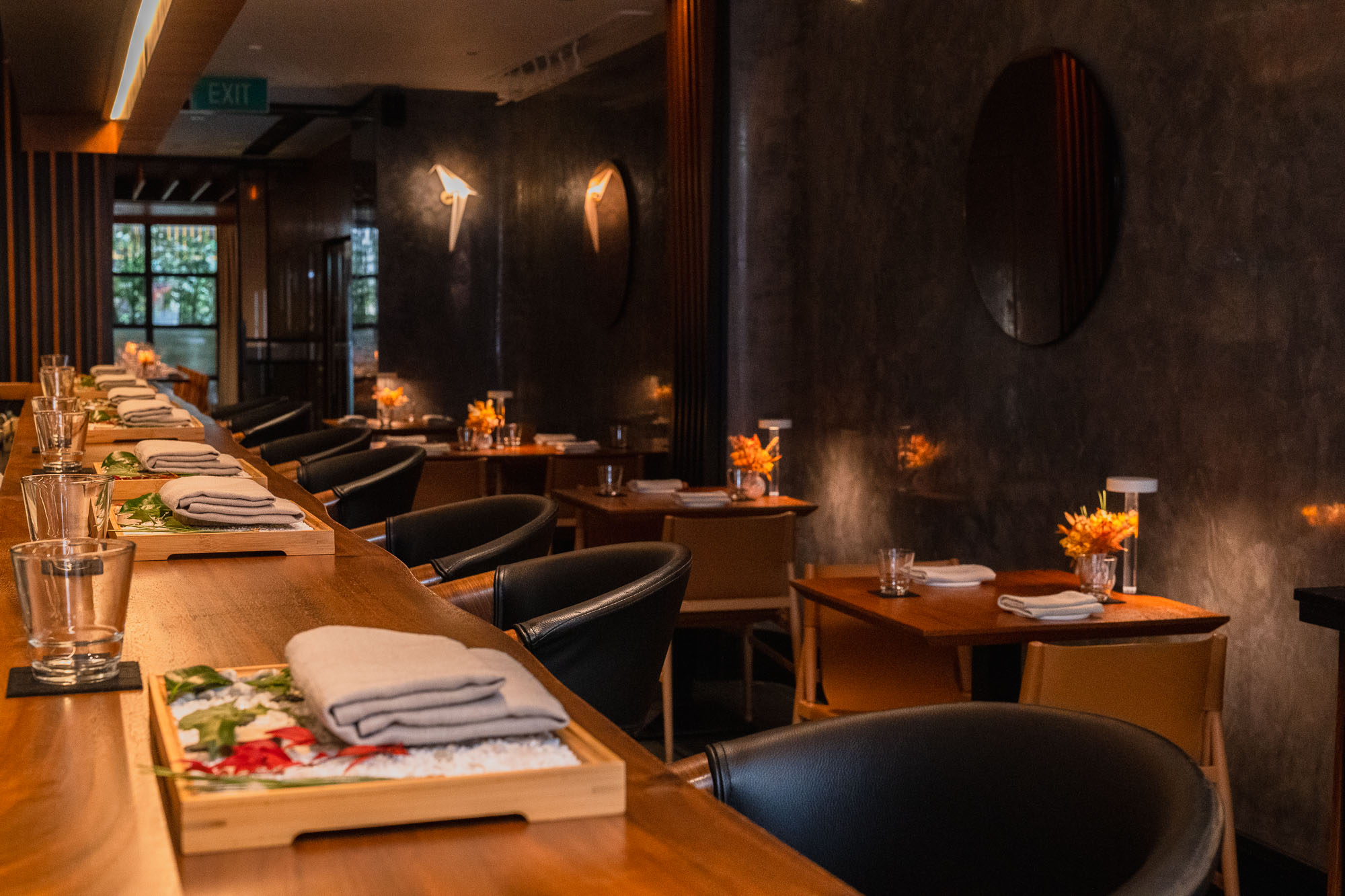 Willow full dining room — long timber omakase counter with black barrel chairs and oshibori trays, dark venetian plaster walls, origami bird sconces and round mirror discs, orange florals