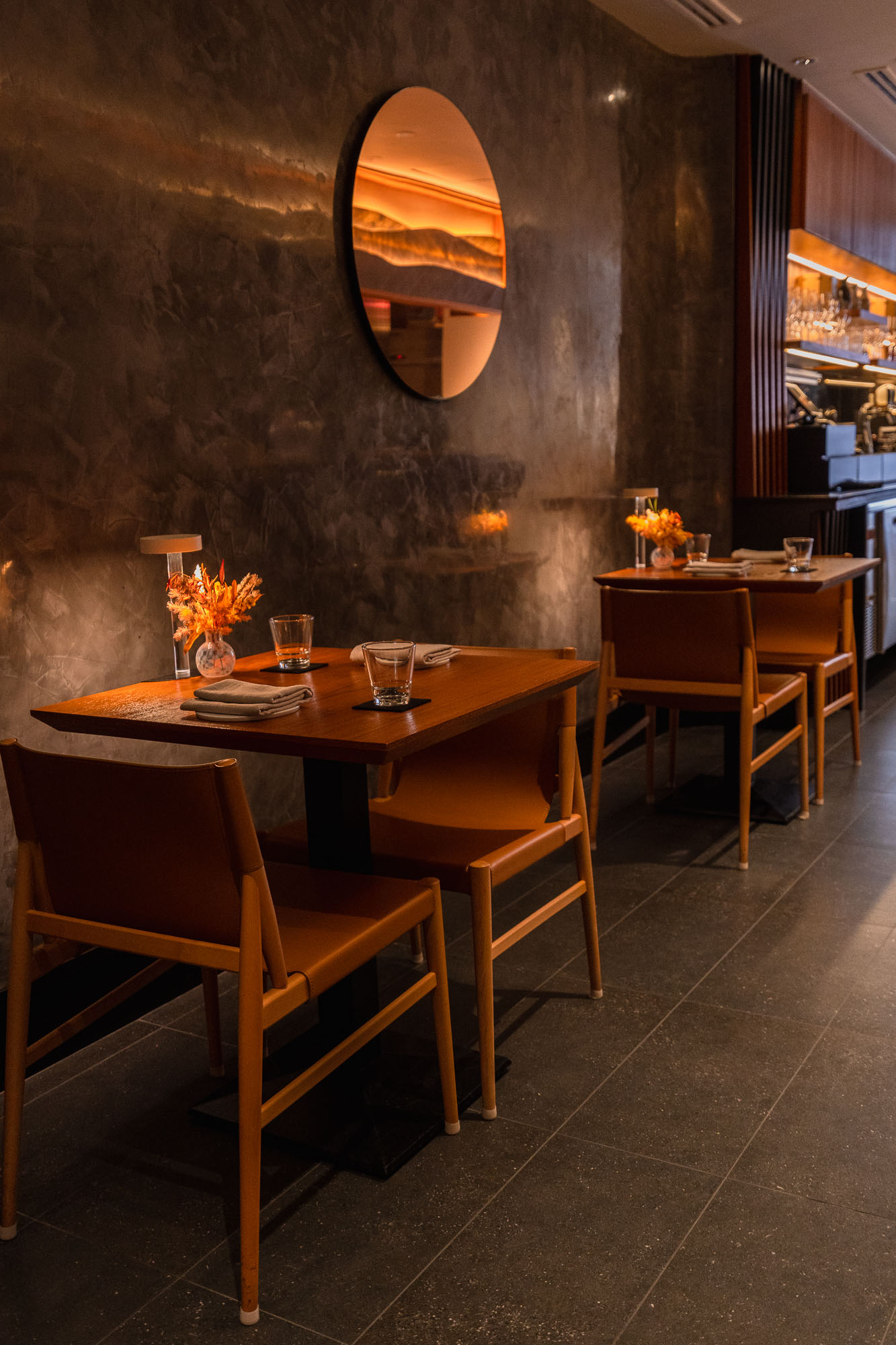 Willow side dining alcove — two dark timber tables with tan leather mid-century chairs, circular amber mirror on dark plaster wall, timber slat bar shelving behind