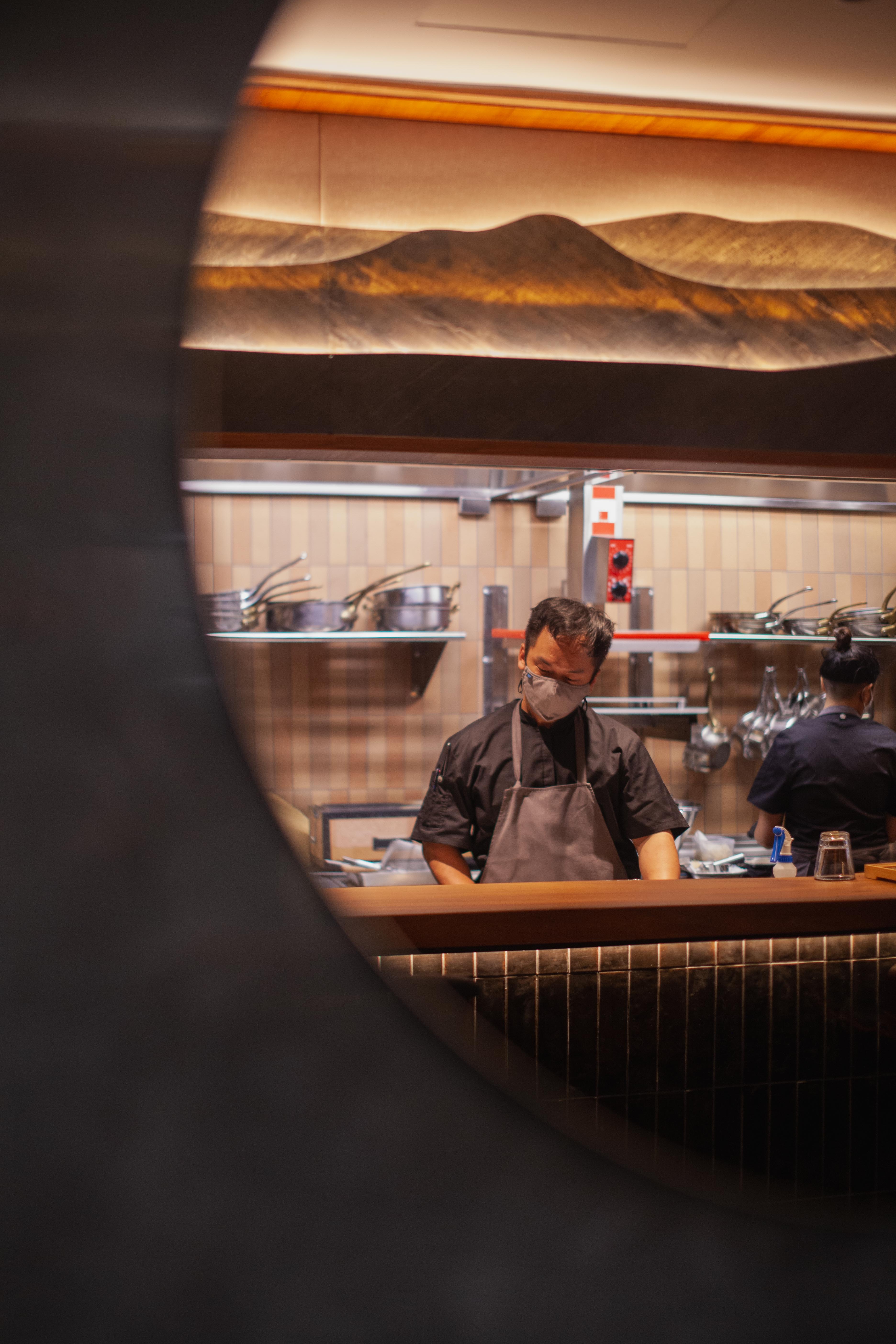 Chef Nicolas Tam framed through a round porthole mirror — kitchen view with mountain sculptural canopy and pink terracotta tile splashback